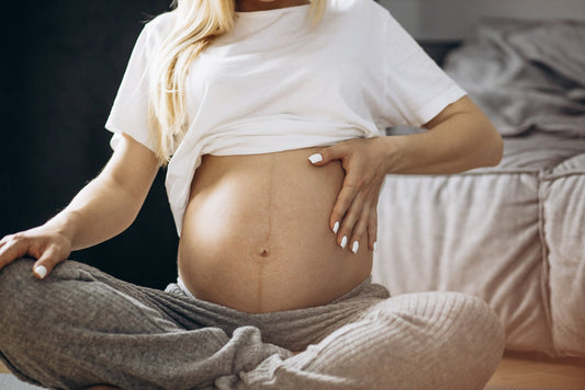A pregnant person sitting on the floor with belly exposed while meditating. She is wearing a white shirt and grey sweatpants.