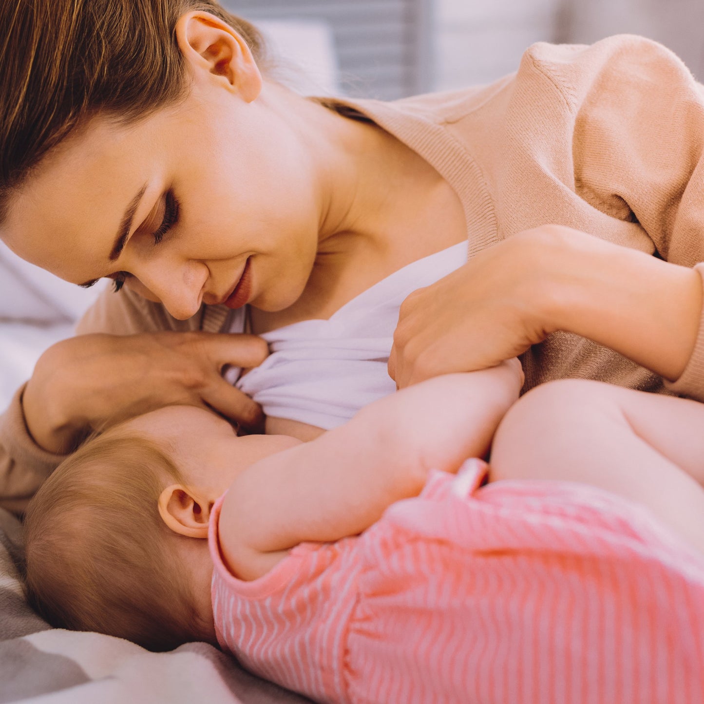 Mom breastfeeding baby in bed in the side lying position. Baby is wearing a pink dress. Mom is wearing a white shirt and beige sweater, while gazing at baby.