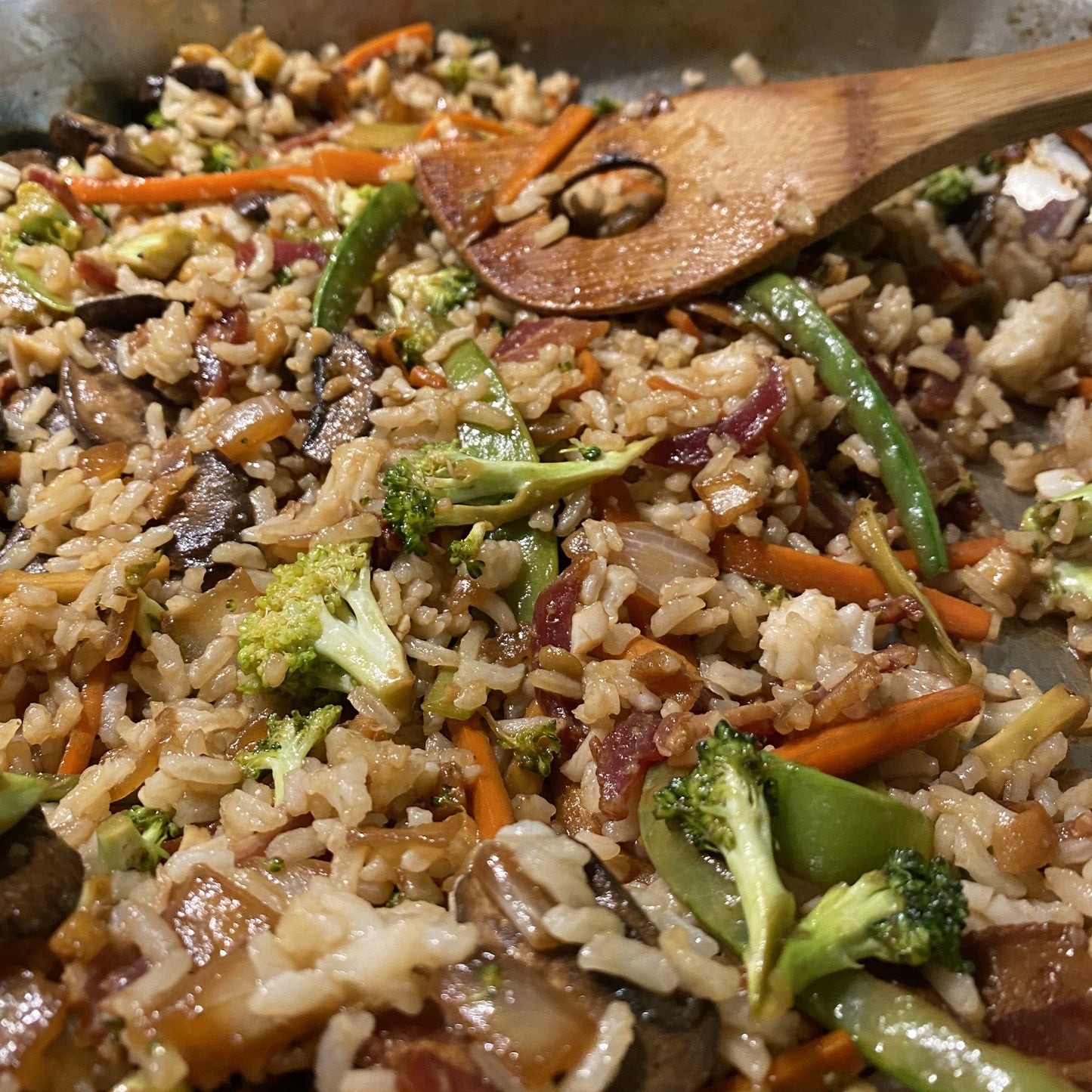 Vegetable stir fry in a pan with a wooden spoon. The stir fry includes broccoli, onion, carrots, mushrooms, bacon, rice, and a scrambled egg.