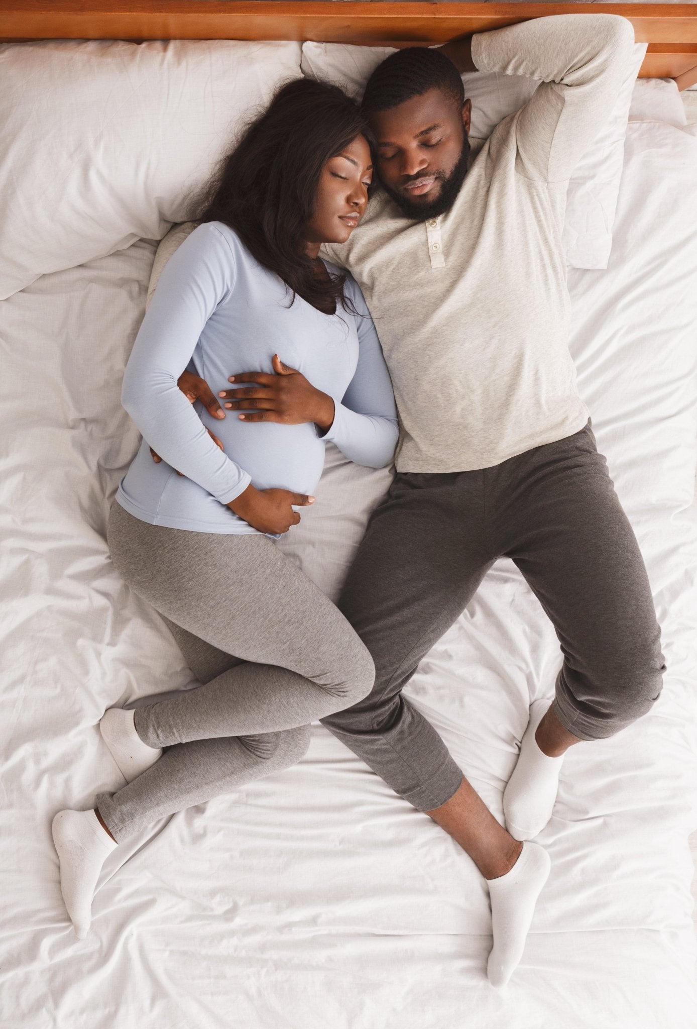 Pregnant woman and partner snuggled in bed on white sheets. Both are napping and look content.
