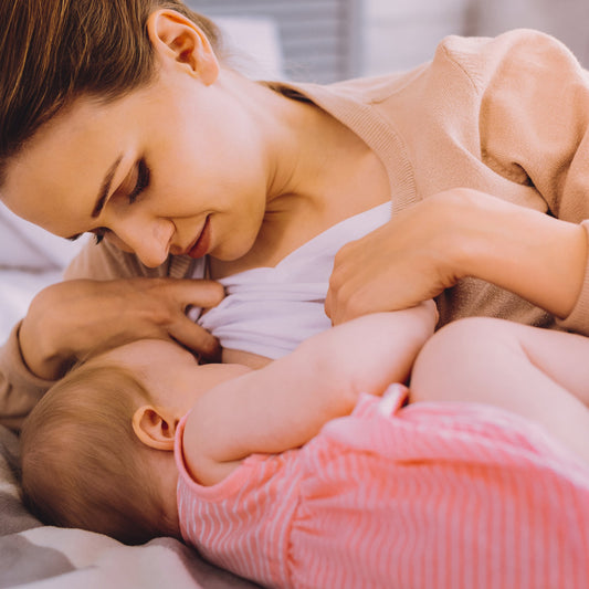 Mom breastfeeding baby in bed in the side lying position.  Baby is wearing a pink dress.  Mom is wearing a white shirt and beige sweater, while gazing at baby.