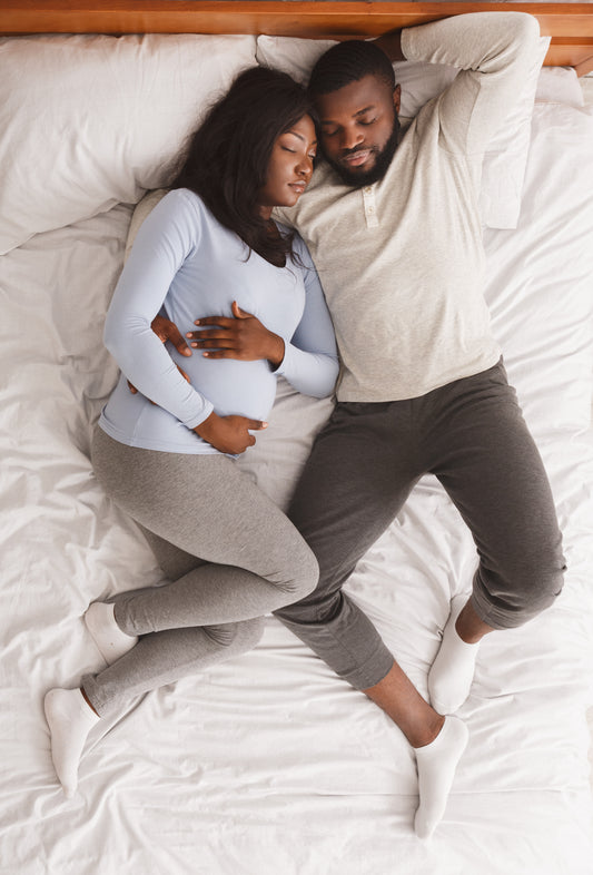 Pregnant woman and partner snuggled in bed on white sheets.  Both are napping and look content.