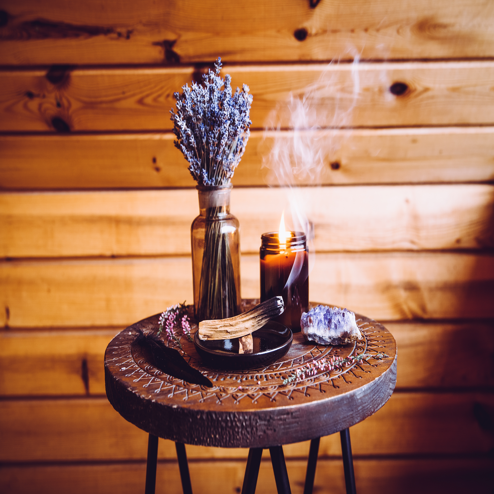 A carved wooden table in a wood panneled room topped with a vase of lavender, burning incense, a lit candle, and a crystal.